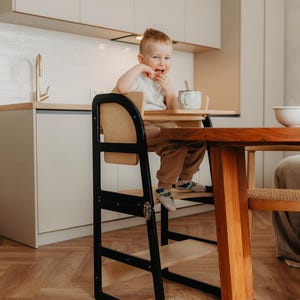 以下が含まれることがあります： 黒と自然な木製の幼児用ステップスツール。子供が木製のテーブルに座っています。ステップスツールは黒いフレームと自然な木製の座席とフットレストを備えています。子供は白いシャツと茶色のズボンを着ています。