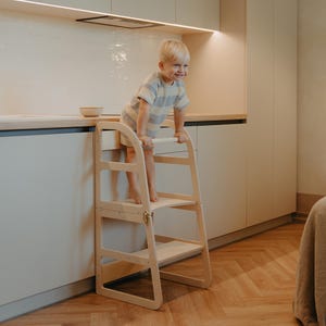 May include: A natural wood step stool with two steps, designed for children. The stool has a simple, modern design with rounded edges and a safety bar at the top. A young child is standing on the top step, smiling. The stool is in a kitchen setting.