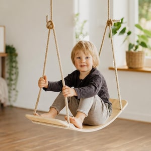 Puede incluir: Un asiento de columpio de madera suspendido por dos cuerdas gruesas de color natural. El asiento es una tabla curva y lisa hecha de madera de tono claro. Las cuerdas están aseguradas a través de orificios a cada lado de la tabla.