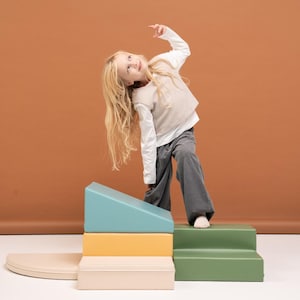 Può includere: Un bambino è in piedi su un set di blocchi di schiuma colorati. I blocchi sono nelle tonalità di blu, giallo, verde e beige. Il bambino indossa una camicia bianca a maniche lunghe, un gilet beige e pantaloni di velluto a coste grigi.
