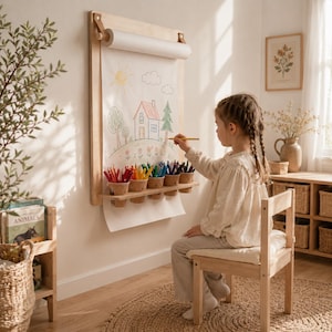 Peut inclure: Un support mural en bois clair pour rouleau de papier, doté d'une étagère horizontale avec cinq petits gobelets remplis de crayons de couleur. Une grande feuille de papier blanc pend du rouleau, montrant un dessin coloré de maison.
