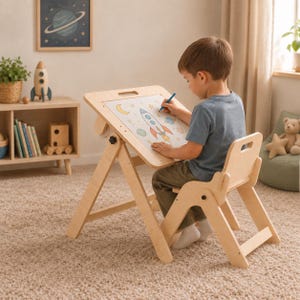 Puede incluir: Un escritorio tipo caballete ajustable de madera clara y una silla a juego. La superficie del escritorio está inclinada y sostiene una hoja de papel con un dibujo colorido de un cohete, estrellas y planetas. La estructura incluye perillas negras.