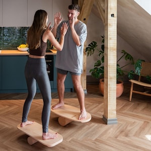 May include: Two light-coloured wooden balance boards with a smooth, oval-shaped top surface and a cylindrical base underneath. Each board is designed for standing on, featuring a natural wood grain finish and a sturdy, minimalist construction.