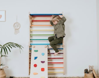 Pode incluir: Um conjunto de escalada em madeira com degraus horizontais num gradiente de cores arco-íris. A estrutura inclui uma rampa amovível com várias pegas de escalada geométricas de cores diferentes fixadas na superfície frontal.