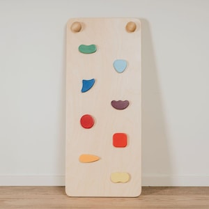 Puede incluir: Un muro de escalada de madera con presas de colores. El muro es de madera clara y presenta presas de escalada de varias formas y colores, incluyendo verde, azul, rojo y amarillo. Dos pomos de madera en la parte superior.