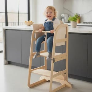 May include: A wooden toddler step stool with a safety bar and footrest. The stool is made of light-colored wood and has a high back and a small seat. A child is sitting in the stool, wearing denim overalls. The stool is designed for use in a kitchen or dining area.