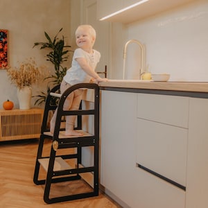 May include: A black wooden step stool with two steps and a safety bar. The step stool is designed for children to reach countertops. The child is standing on the top step, smiling, and reaching for the countertop.