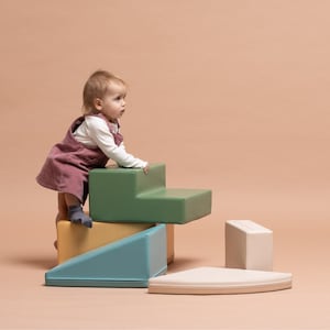 Può includere: Un bambino con un vestito rosa e una camicia bianca a maniche lunghe gioca su un set di blocchi di schiuma colorati. I blocchi sono di varie forme e colori, tra cui verde, giallo, blu e beige. Lo sfondo è di colore pesca uniforme.