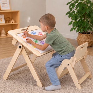 May include: A light-colored wooden easel desk with an adjustable slanted top and a matching wooden chair. The desk surface holds a large sheet of white paper featuring colorful stamped paw prints in blue, red, green, and purple ink.
