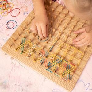 May include: A wooden pegboard with colorful rubber bands being used to create geometric patterns. The board has numerous wooden pegs arranged in a grid. A child's hands are shown manipulating the rubber bands, creating shapes with yellow, blue, red, and white bands.