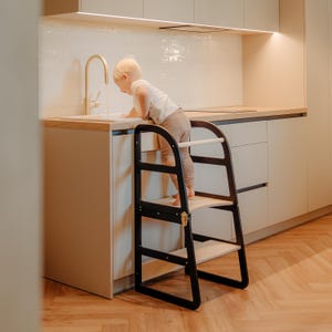 May include: A black and natural wood kitchen step stool with two steps. The step stool has a curved design and a safety bar. A child is standing on the top step, reaching towards a sink.