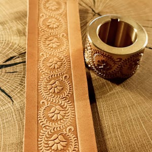 May include: A close-up of a tan leather strip with an embossed floral design. A brass tool with a matching floral pattern sits next to the leather. The tool is cylindrical with an open centre.