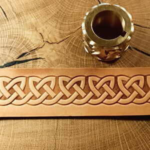 May include: A brass object with a circular design and a light brown leather strip with a Celtic knot pattern. The objects are on a wooden surface. The leather strip is approximately 3 cm wide.