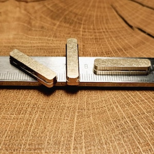 Op de afbeelding: Close-up van drie kleine, rechthoekige, goudkleurige metalen objecten. Eén staat rechtop, een ander ligt plat en de derde is schuin. Ze zijn geplaatst op een metalen liniaal met het cijfer 8 zichtbaar, tegen een houten achtergrond.