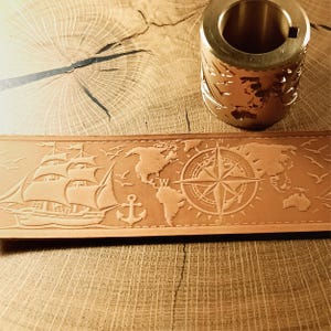 Puede incluir: Un cinturón de cuero marrón claro con temática náutica, con un diseño en relieve de un velero, ancla, brújula y mapa del mundo. Un cilindro de metal dorado con un diseño de mapa del mundo está en el fondo. El cinturón está sobre una superficie de madera.