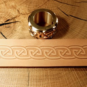 May include: A brass leather stamping tool and a strip of stamped leather. The tool is cylindrical with a decorative Celtic knot design. The leather strip displays a repeating Celtic knot pattern, showcasing the tool's impression.