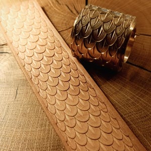 May include: A close-up of a leather craft tool and a strip of leather. The tool is a brass roller with a repeating scale pattern. The leather strip is light brown and has the same scale pattern embossed on it.