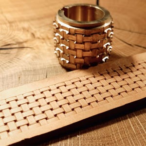 Op de afbeelding: Een messing leder stempelgereedschap met een geweven patroon. Het gereedschap ligt op een stuk leer dat met hetzelfde patroon is gestempeld.