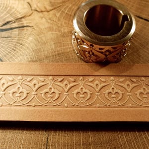 Op de afbeelding: Een close-up van een messing embossing tool met een sierlijk bloemenontwerp. Een strook bruin leer met een reliëf bloemenpatroon ligt eronder, op een houten oppervlak.