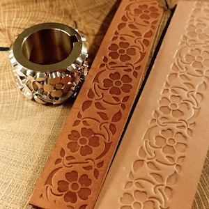 May include: A metal floral design stamp and two leather strips with floral patterns. One strip is stamped with the design, and the other is embossed. The leather strips are shades of brown. The stamp is a cylinder with a floral pattern.