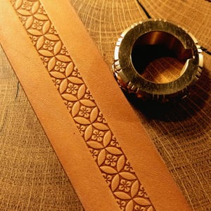May include: A tan leather strip featuring a continuous embossed pattern of interlocking circles with small floral centres, positioned next to a circular metallic tool with a notched edge and a central opening used for leather stamping.
