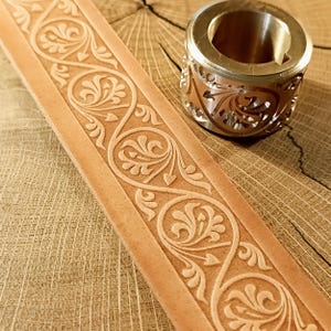 Puede incluir: Primer plano de una tira de cuero marrón claro con un diseño floral en relieve. Un anillo de metal con un patrón floral a juego se encuentra junto al cuero. El anillo tiene un exterior dorado y un interior hueco.