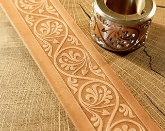 May include: Close-up of a light brown leather strip with an embossed floral design. A metal ring with a matching floral pattern sits next to the leather. The ring has a gold-colored exterior and a hollow interior.