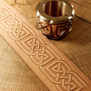 May include: A close-up of a light brown leather strap with an embossed Celtic knot design. A gold-colored metal ring with a matching Celtic knot pattern sits beside the strap on a wooden surface.