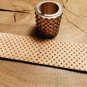 Puede incluir: Un primer plano de una herramienta de estampado de cuero con un patrón tejido. La herramienta está hecha de metal y tiene una forma circular. La herramienta está sobre una superficie de madera.