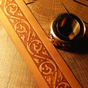 May include: A tan leather strip featuring a continuous embossed scrollwork pattern alongside a metallic ring with a matching cut-out design. The leather has a smooth finish with darker brown recessed areas highlighting the intricate floral-like motifs.