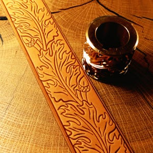 Pode incluir: Uma tira de couro bege com um padrão profundamente gravado de folhas de carvalho e pequenas bolotas, posicionada ao lado de uma ferramenta de estampagem cilíndrica de metal com um design de folha intrincado na superfície.