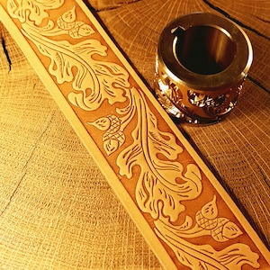 May include: A tan leather strip featuring a deeply embossed pattern of oak leaves and acorns, positioned next to a cylindrical metal stamping tool with a hollow centre and a keyway slot, used for creating detailed leather impressions.
