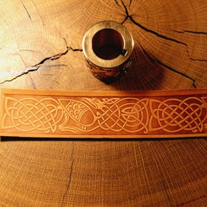 Puede incluir: Una tira de cuero color canela con un patrón en relieve de una figura de ave compleja integrada en diseños de nudos continuos, colocada debajo de un objeto metálico cilíndrico con una abertura circular y una pequeña muesca lateral.