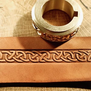 Puede incluir: Primer plano de un anillo de latón con un diseño calado decorativo, apoyado sobre una correa de cuero marrón con un patrón de nudo celta en relieve. La correa de cuero es de un color marrón cálido y rico.