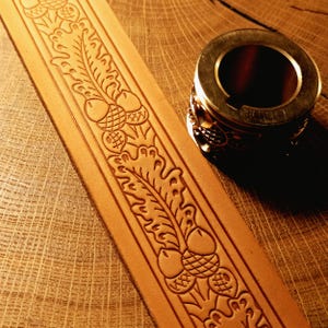 May include: A light tan leather strip featuring a stamped pattern of oak leaves and acorns, positioned next to a cylindrical metal stamping tool with a matching engraved design on its side, showing the tool used for leathercrafting impressions.