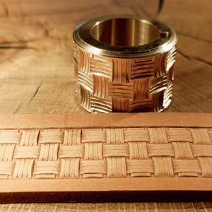 Op de afbeelding: Een messing leerstempelgereedschap met een mandweefpatroon. De tool is cilindrisch met een gedetailleerd geweven ontwerp. Een leren strook met een bijpassend mandweefpatroon staat op de voorgrond.
