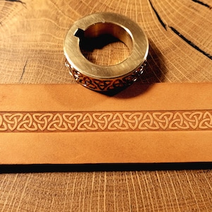 May include: A brass ring with an openwork Celtic knot design rests on a light brown leather belt featuring an embossed Celtic knot pattern. The items are displayed on a wooden surface.