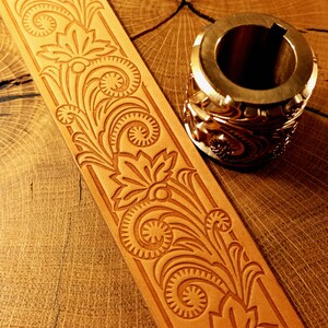 May include: A tan leather strip featuring a deeply embossed floral scroll pattern alongside a cylindrical metal stamping tool. The tool displays a matching intricate botanical design on its exterior surface, designed for creating impressions on leather.
