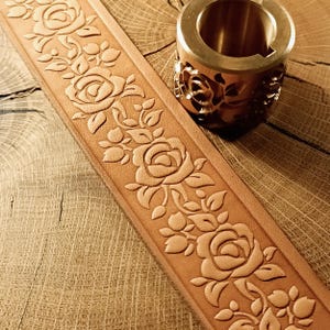 Op de afbeelding: Close-up van een bruinleren strook met een reliëfpatroon van rozen en bladeren. Een messing metalen gereedschap met een rozendesign ligt naast het leer. De items liggen op een houten oppervlak.
