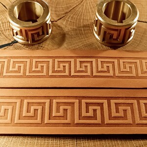 Könnte beinhalten: Zwei Messingringe mit geometrischem Design liegen über zwei Streifen braunen Leders, die mit einem griechischen Schlüsselmuster geprägt sind. Die Artikel werden auf einer Holzoberfläche präsentiert.
