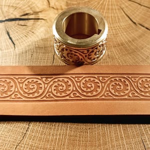 May include: A brass metal ring with an ornate, openwork design sits atop a piece of brown leather with a matching embossed pattern. The leather strip displays a repeating swirl and scroll design. The background is a wooden surface.