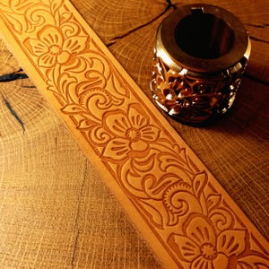May include: A light tan leather strip featuring a continuous embossed floral pattern with swirling vines and petals, positioned next to a cylindrical metal stamping tool that displays a matching intricate floral design on its exterior surface.