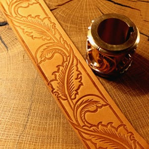 Puede incluir: Una tira de cuero color canela con un patrón de hojas florales en relieve continuo, junto a una herramienta de estampado cilíndrica de metal con un diseño de hoja grabado en su superficie exterior para marcar cuero.