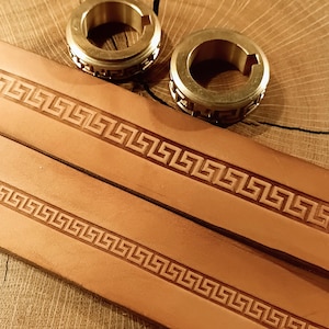 Könnte beinhalten: Nahaufnahme von zwei Messingringen und zwei hellbraunen Lederriemen. Die Lederriemen weisen ein geprägtes griechisches Schlüsselmuster auf. Die kreisförmigen Ringe haben ein dekoratives geometrisches Design.