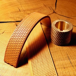 May include: A tan leather strip featuring a repeating diamond-shaped embossed pattern and a cylindrical brass knurling tool with a matching diamond-textured surface. Both items exhibit a consistent geometric design and metallic or natural tan tones.