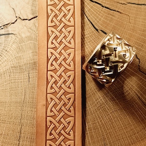 Puede incluir: Primer plano de una tira de cuero marrón claro con un patrón de nudo celta en relieve. A la derecha, un sello de metal dorado con un diseño similar. El cuero y el sello se encuentran sobre una superficie de madera.