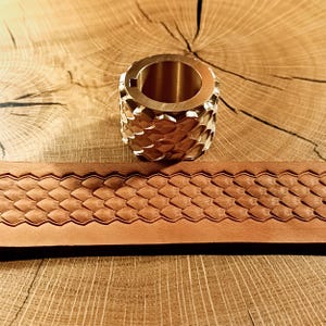 Puede incluir: Un anillo de latón con un patrón de escamas y un cinturón de cuero marrón con un diseño de escamas a juego se muestran sobre una superficie de madera. El anillo tiene forma cilíndrica.
