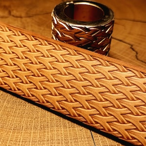 Puede incluir: Una tira de cuero color canela con un patrón de tejido de cesta repetitivo grabado en su superficie, colocada junto a una herramienta de estampado cilíndrica de metal con un diseño texturizado a juego grabado en su borde exterior.