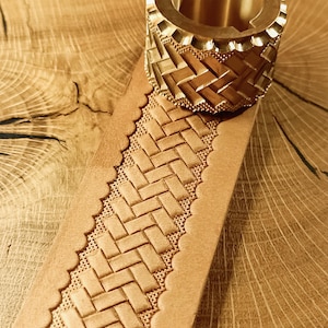 Op de afbeelding: Close-up van een lederbewerkingstool van messing en een stuk bruin leer. De tool heeft een patroon. Het leer is voorzien van een reliëf geweven patroon en heeft een geschulpte rand. De achtergrond is een houten oppervlak.
