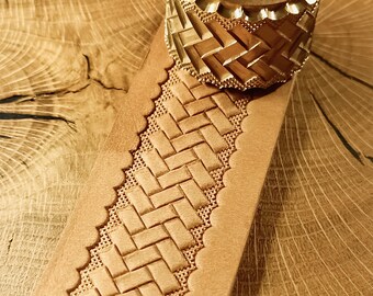May include: Close-up of a leather craft tool and a piece of tan leather. The tool is made of brass and has a patterned design. The leather has a woven pattern embossed into it, with a scalloped edge. The background is a wooden surface.
